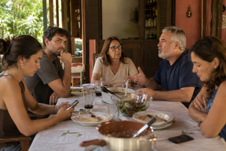 Família brasileira em almoço de domingo com clima tenso após discussão política, enquanto alguns olham para o celular e outros evitam contato visual.